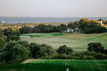 Malaga, Spain - January, 21, 2024 -  Overlooking a golf course green with golfers, a cart, and a backdrop of the sea and trees at dusk.
