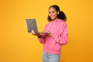 Cheerful black youngster girl with laptop browsing for homework, studio