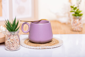 An elegant scene: a purple coffee cup with a golden spoon beside a green plant in a glass jar, set on a white table amidst boho decor. Ideal for café banners. 
