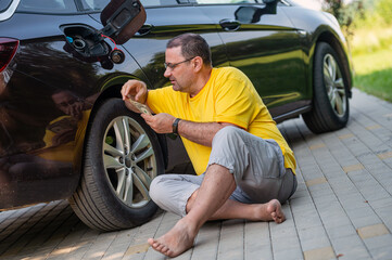 worried man sitting on the pavement next to a car near an open fuel tank, fuel price rise concept