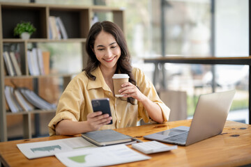 Attractive Asian businesswoman sitting and relaxing with a hot drink, using the mobile phone at the desk in the office