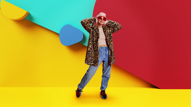 Beautiful young smiling girl in stylish in leopard print coat and red sunglasses listening to music in headphones against multicolored background. Fashion brand ads. Concept of youth, lifestyle