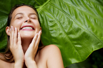 Organic skin care. A joyful woman with glowing skin touches her face gently, her eyes closed with a wide smile, behind a large green leaf.