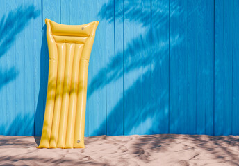 3D rendering of a yellow inflatable air mattress leaning against a vibrant blue wooden wall on sandy ground with palm tree shadows, beach vacation concept.