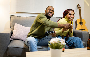 A dark-skinned couple is sitting on the couch at home playing video game consoles with wireless...