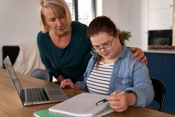 Down syndrome woman studying on computer and notebook with help of her mother