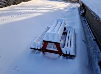 a camp bench with a table covered in a layer of snow. a child sweeps them with a snow shovel....
