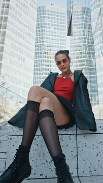 Vertical shot seductive woman wearing black jacket short skirt stockings and high boots sitting on stairs playfully shaking leg near glass skyscrapers modern buildings. Urban style fashion model