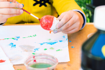 11-year-old boy watercolour painting an Easter egg with a paint brush on a table in his backyard.