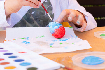 11-year-old boy watercolour painting a blue and red Easter egg with a paint brush on a table in his backyard.