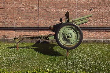 Old green metal cannon from Second World War with red brick wall in background.
