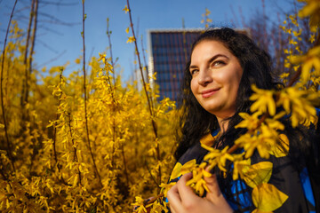 Young happy smiling woman plus size in spring in city park among blooming yellow forsythia bushes
