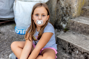 Photo of 4 years old girl looking into camera with a dummy in her mouth. Girl acting like baby.