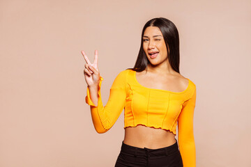 Young latin american happy brunette woman does peace victory sign with hand and winks eye. Studio portrait isolated over beige background.