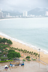 Public beach park sandy shoreline surrounding by lush green tropical trees, concrete square, row of high-rise hotels, apartment, office building, mountain range background downtown Nha Trang