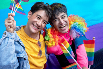 Two gay men sitting on rainbow flags on the ground, celebrate by waving flags to welcome the pride parades during Pride Month celebrations, LGBTQ concept