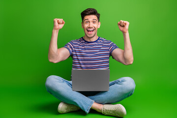 Full length photo of impressed man wear stylish t-shirt sitting on floor with laptop on legs win bet isolated on green color background
