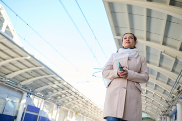 View from the bottom woman commuter holding mobile phone, waiting to board the train, on the platform of railway station