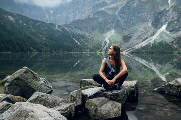 Beautiful young brunette woman in stylish outfit sitting on the rocks, stones on the shore and admiring beautiful view of green hills and mountains on Morskie Oko lake, High Tatras, Zakopane, Poland - obrazy, fototapety, plakaty