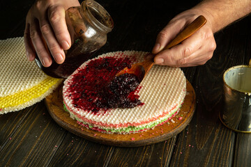 The cook, with a spoon in his hand, spreads jam on a round waffle cake on the kitchen table. Making...