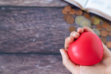 Hand holding red heart over a wooden table with open holy bible book and coins. Christian biblical concept of tithing, giving, sharing, thanksgiving, and generosity.