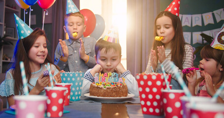 Portrait of sad birthday boy sitting in living room near cake with friends blowing party whistles....
