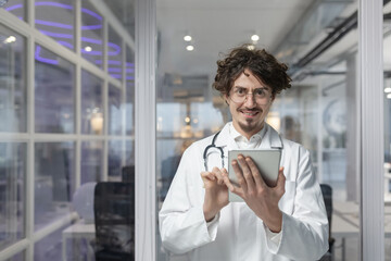 A doctor in a white medical coat and stethoscope holds a tablet for a medical consult in his office. Smile man looking at camera