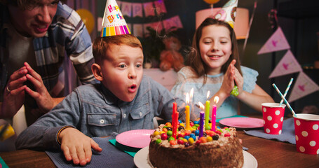 Happy red hair boy in a festive cap blows out the colorful candles cake on birthday party, family...