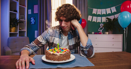 Alone Caucasian upset guy sitting at the birthday cake and looking with sad eyes on it, then...