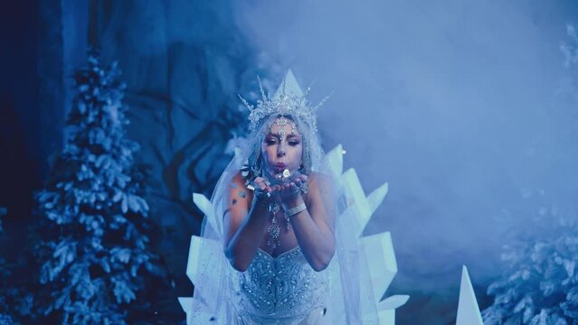 Art portrait Fantasy woman snow queen blows on shining sparkles glitter creating magic cold in forest, white dress crown icicles. Fairy tale girl frozen princess lady Winter nature trees ice blue fog