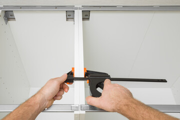 Young adult man hands using f speed clamp and pressing white boards of base cabinet frame....
