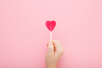 Little child hand fingers holding heart shape lollipop on stick on light pink table background. Pastel color. Sweet candy. Closeup. Top down view. - obrazy, fototapety, plakaty