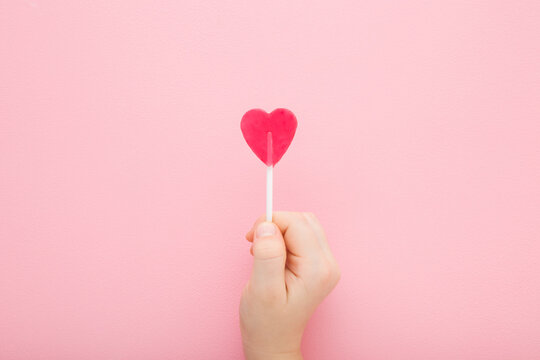 Little child hand fingers holding heart shape lollipop on stick on light pink table background. Pastel color. Sweet candy. Closeup. Top down view.