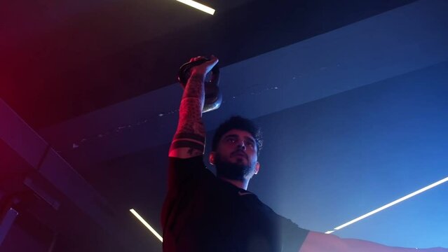 A man performs a kettlebell snatch in a gym, with artistic blue and red lighting and a subtly misty atmosphere