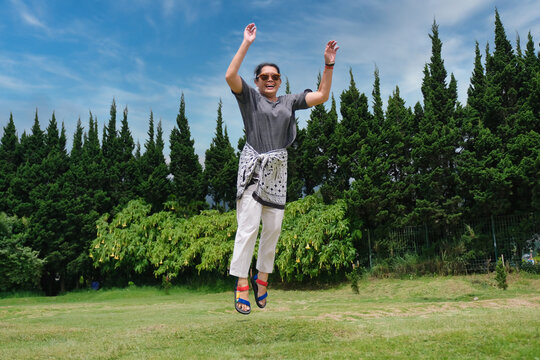 A woman wearing sunglasses is in the park, celebrating the sunny day by jumping as high as she can