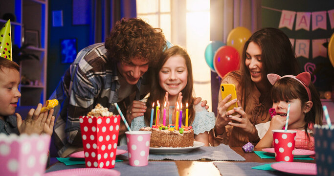 Caucasian pretty birthday girl in conus sitting at the table with friends while father or older brother coming and bringing birthday cake.