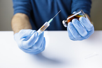 The hands of the doctor or nurse, on a white table and wearing blue gloves, hold a syringe with a...