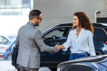 Happy salesman congratulating his female customer for buying a new car in a showroom. Salesperson...
