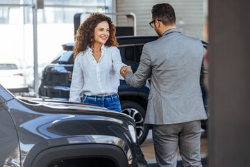 Happy salesman congratulating his female customer for buying a new car in a showroom. Salesperson selling cars at car dealership. - obrazy, fototapety, plakaty