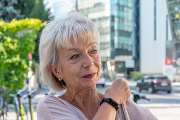 An elderly woman with a modern wristwatch adjusts a bag on her shoulder against a background of modern buildings.