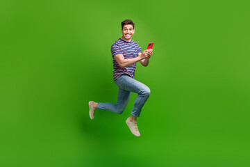 Fototapeta na wymiar Full length photo of funky eccentric man wear stylish t-shirt jeans trousers jumping holding smartphone isolated on green color background