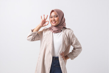 Portrait of excited Asian hijab woman in casual suit showing ok hand gesture and smiling looking at...