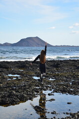 Young woman with long flowing brown hair walking on the rocky Fuerteventura beach in the morning. In the background, the uninhabited island of Lobos and the Lanzarote and the waving Atlantic ocean.