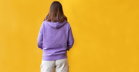 Back view of young woman wearing lilac purple hoodie or hooded sweatshirt against bright yellow wall background mockup friendly copy space