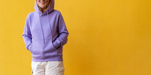 Young happy smiling woman in hood in purple violet hoodie on yellow wall background copy space...