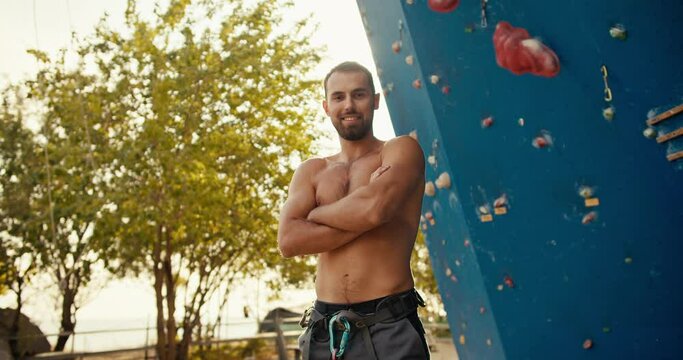 Portrait of a happy brunette guy with a beard who folds his arms on his chest and looks at the camera near a blue climbing wall on a sunny summer day. Experienced man Scala with bare torso posing and