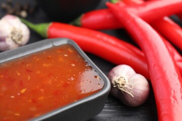 Spicy chili sauce in bowl, garlic and peppers on table, closeup