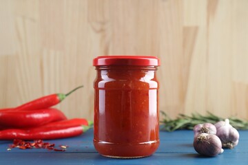 Spicy chili sauce in jar, garlic and peppers on blue wooden table