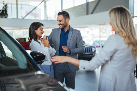 Car Dealer Selling Auto To Young Couple. Discussing The Purchase. Young Family Speaking With Professional Car Seller In Dealership Center