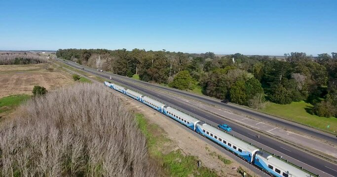 Blue passenger train moving along tracks beside a highway, rural landscape on the outskirts of Buenos Aires.